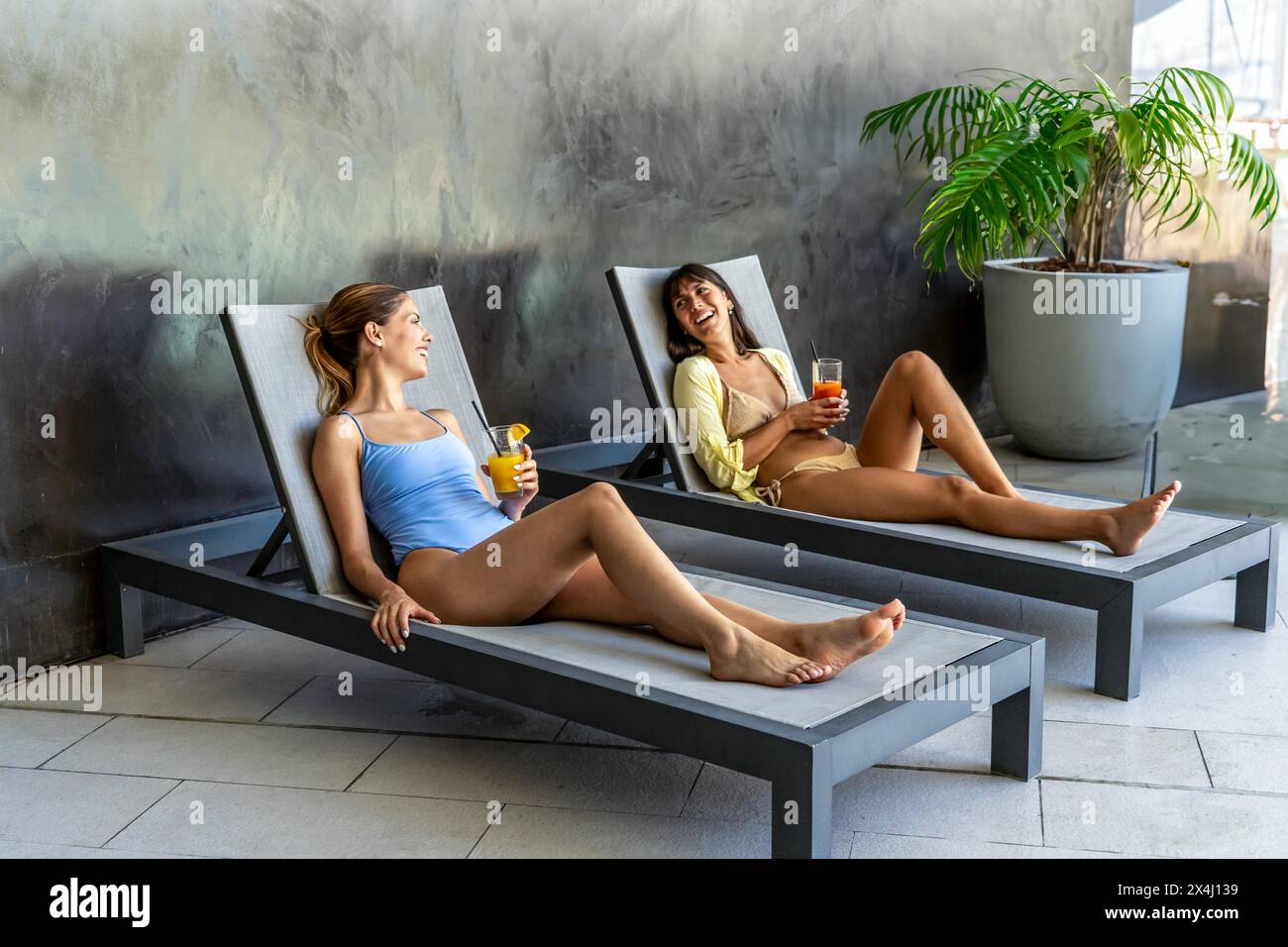 Due amiche stanno ridendo e bevendo succo di frutta nella zona piscina di un hotel elegante Foto Stock