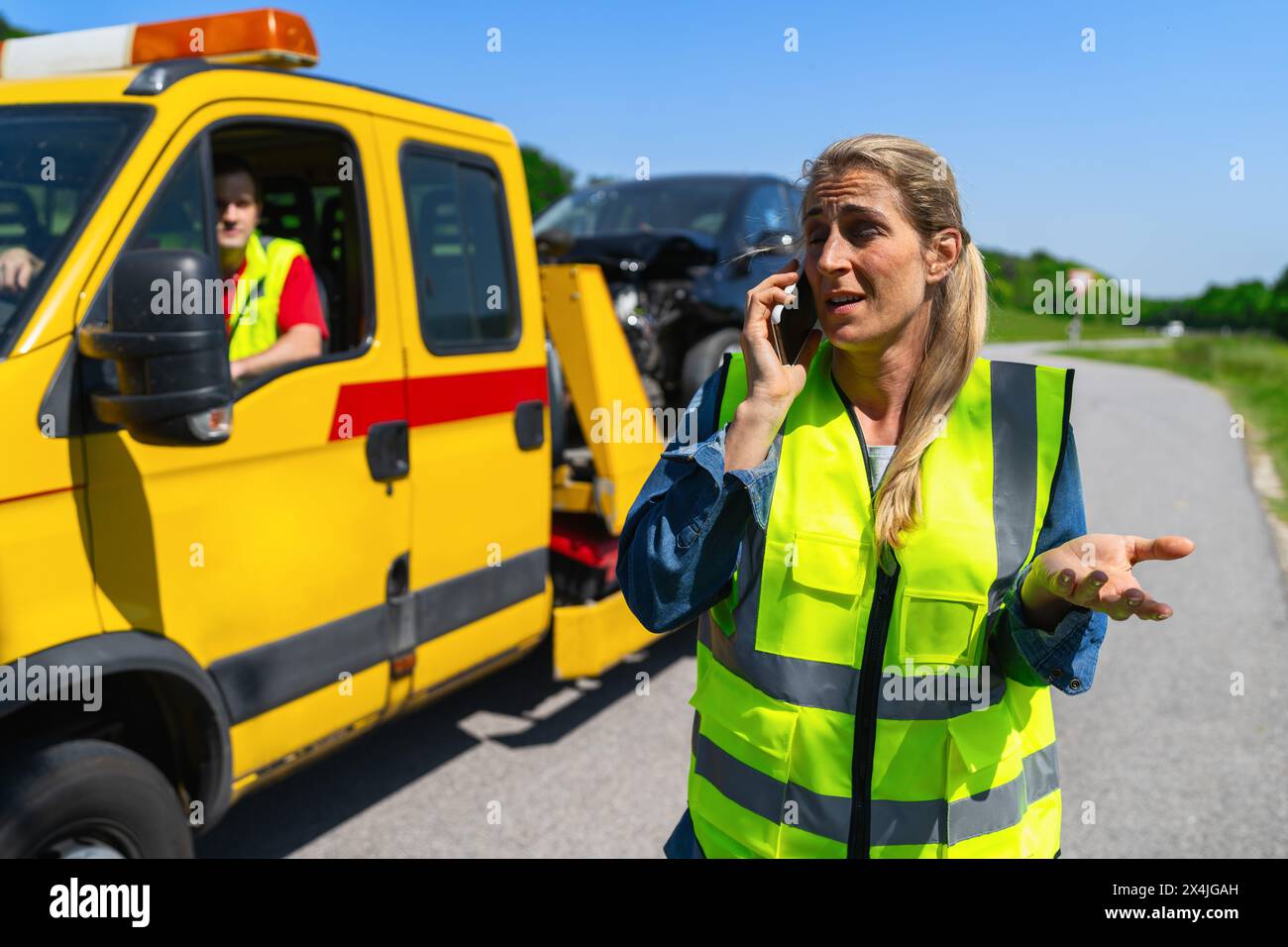 Una donna automobilista gesticolata e parla al telefono dopo che il servizio di traino ha caricato la sua auto su un camion di traino e spiega il suo problema. A lato strada Foto Stock