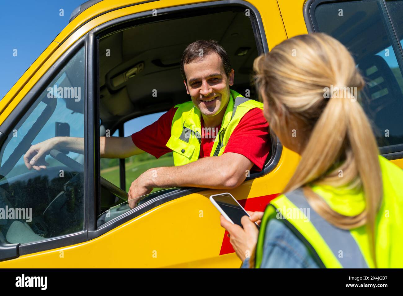 Donna che utilizza lo smartphone che parla con un conducente sorridente di un rimorchio seduto su un camion giallo. Immagine del concetto di assistenza stradale. Foto Stock