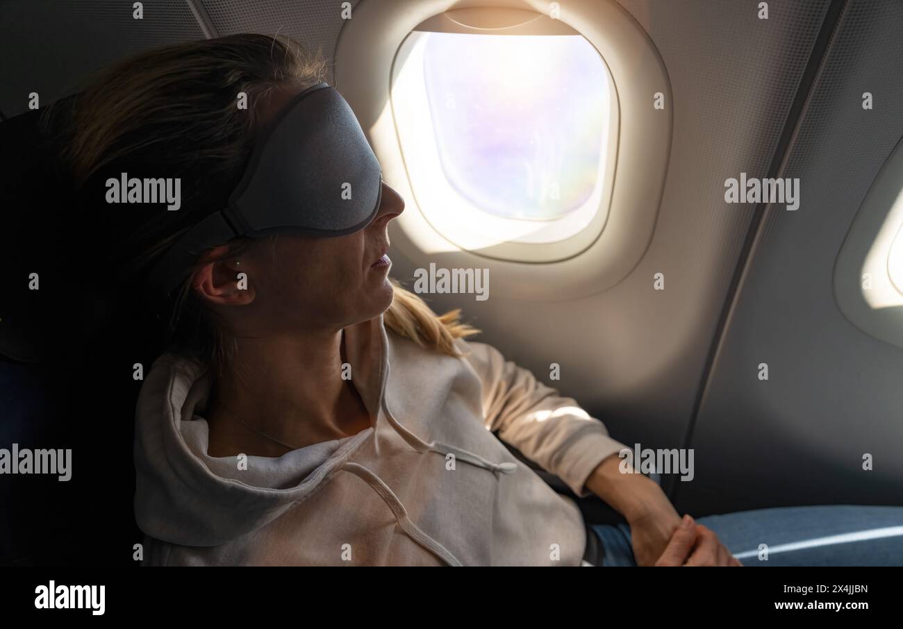 Donna con maschera dormiente sull'aereo appoggiata allo schienale, illuminata dalla luce della finestra Foto Stock