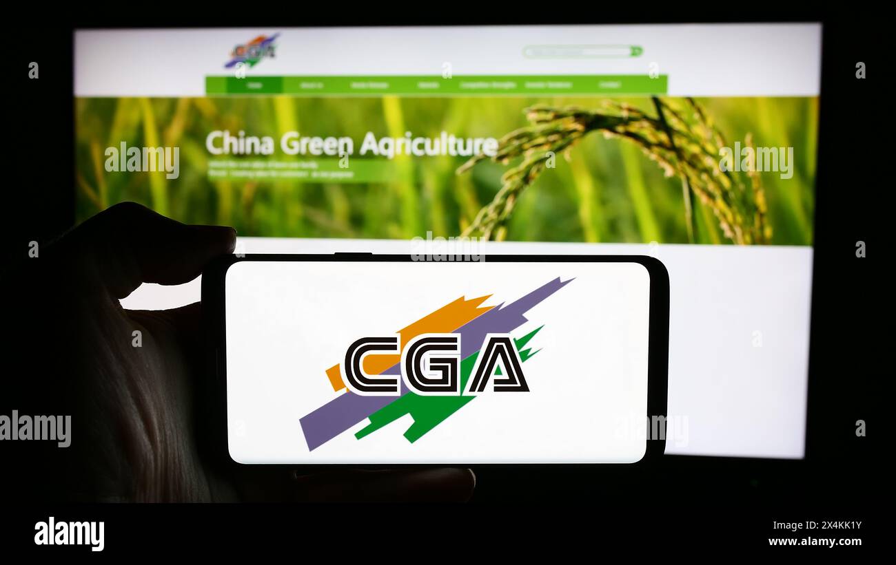 Persona che detiene un cellulare con il logo della società cinese China Green Agriculture Inc. (CGA) di fronte alla pagina Web aziendale. Mettere a fuoco il display del telefono. Foto Stock