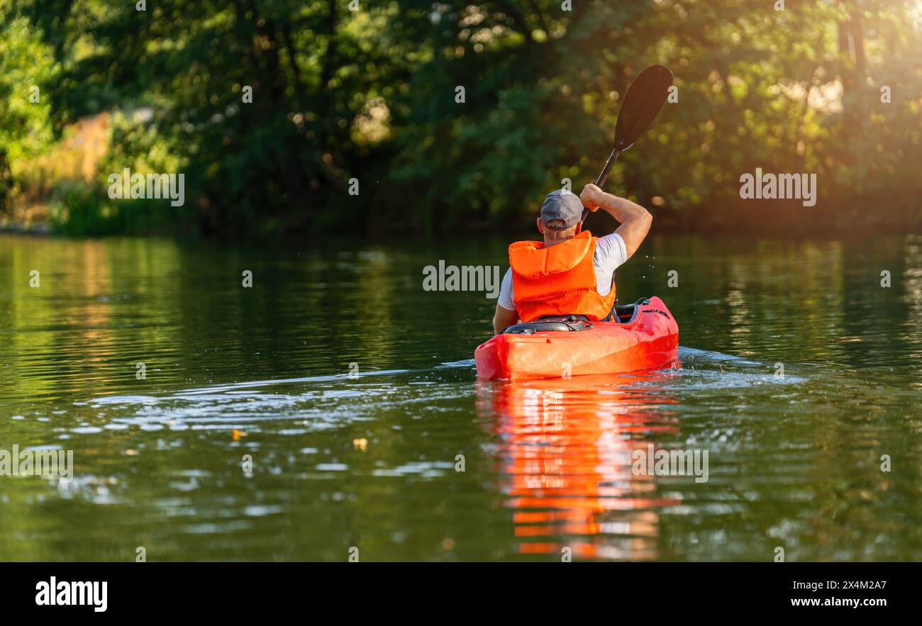 Uomo in un giubbotto salvagente arancione che pagaia un kayak rosso su un fiume calmo, circondato da una vegetazione lussureggiante alla luce del sole. Immagine del concetto di kayak Water Sports Foto Stock