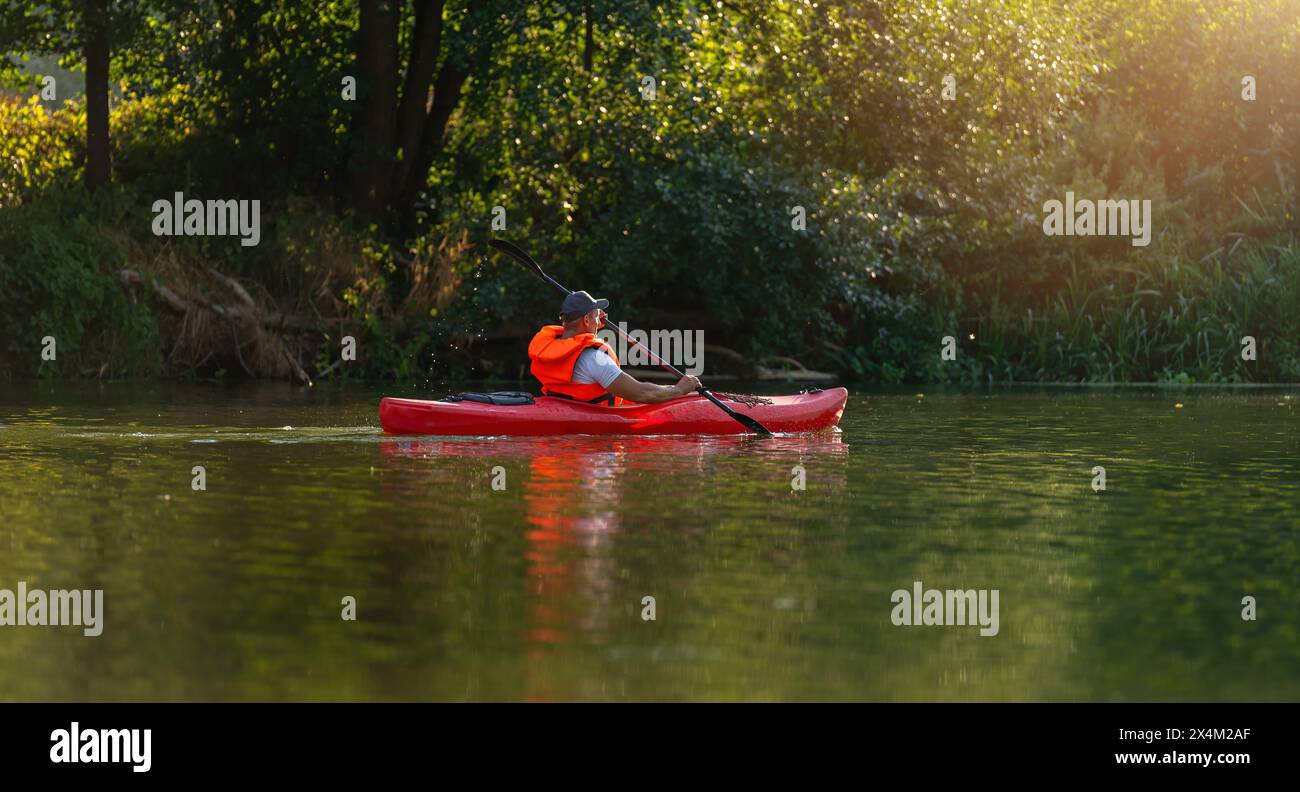 L'uomo a bordo di un kayak rosso pagaia serenamente su un fiume calmo, circondato da fitte foglie verdi sotto la luce del sole dorato in estate. Kayak Sport acquatici concetto i Foto Stock