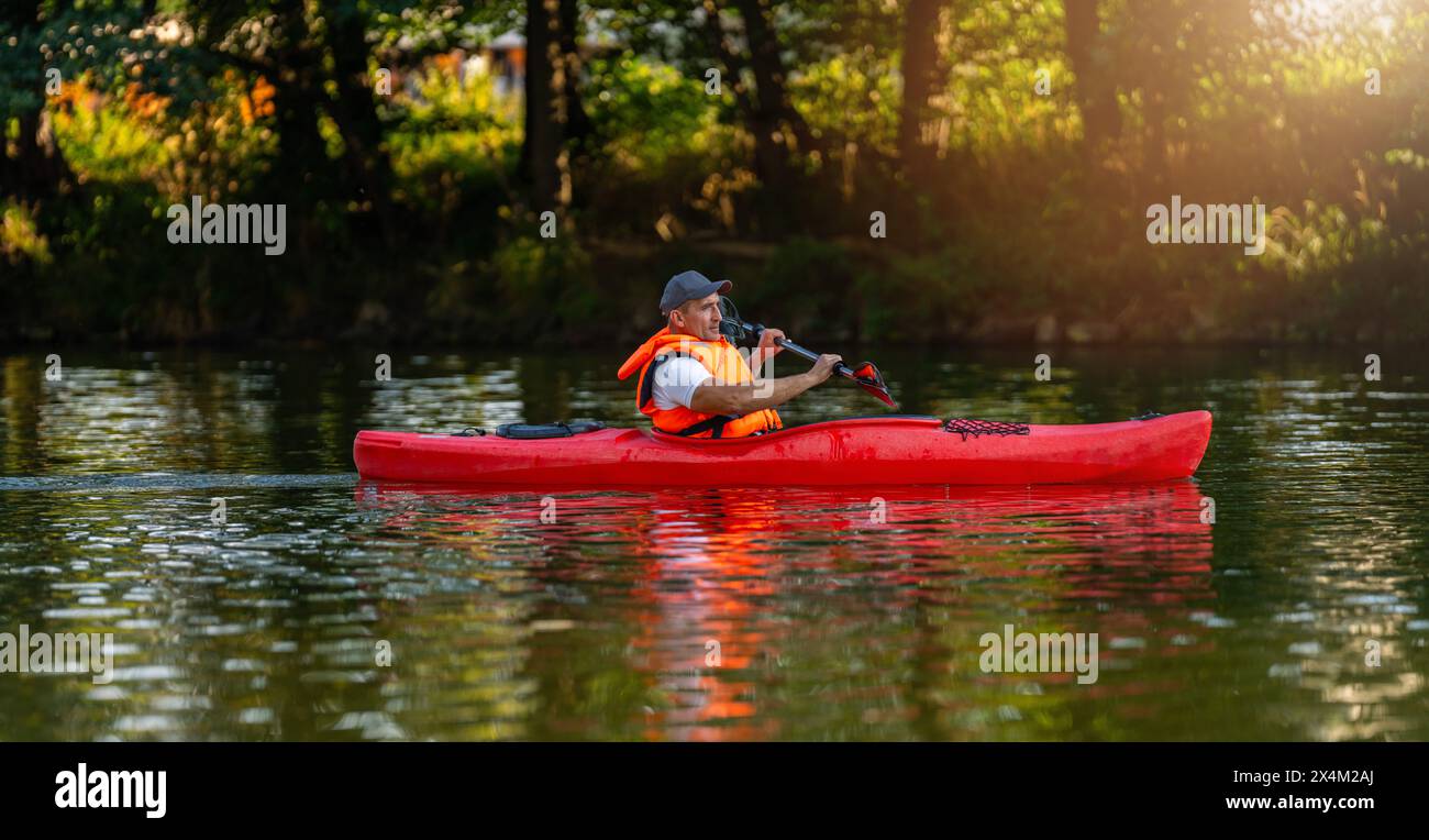 Un uomo che fa kayak da solo in un kayak rosso su un fiume tranquillo, circondato da una vegetazione lussureggiante e dalle luci della sera. Immagine del concetto di kayak Water Sports Foto Stock