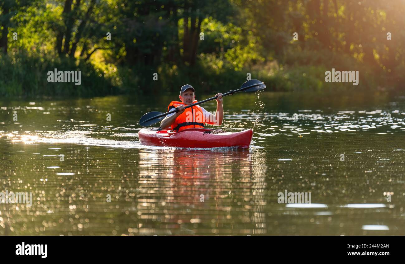 Uomo con giubbotto salvagente arancione che fa kayak rosso su un fiume, luce del sole dorata che si riflette sull'acqua. Immagine del concetto di kayak Water Sports Foto Stock