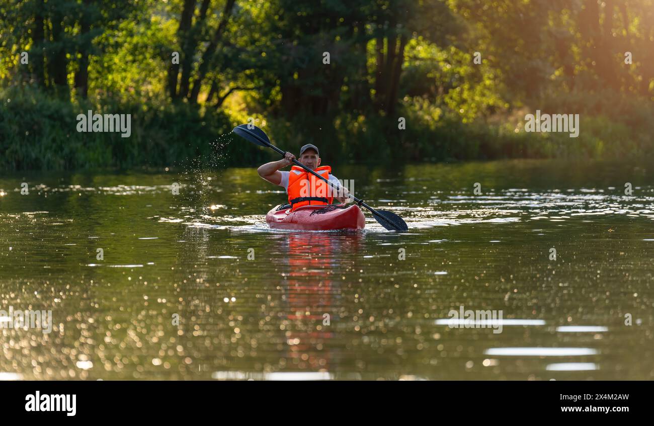 Uomo con giubbotto salvagente arancione, pagaia su un kayak rosso attraverso le calme acque del fiume, circondato dalla luce del sole e circondato da lussureggianti vegetazioni in baviera germania. Kaya Foto Stock