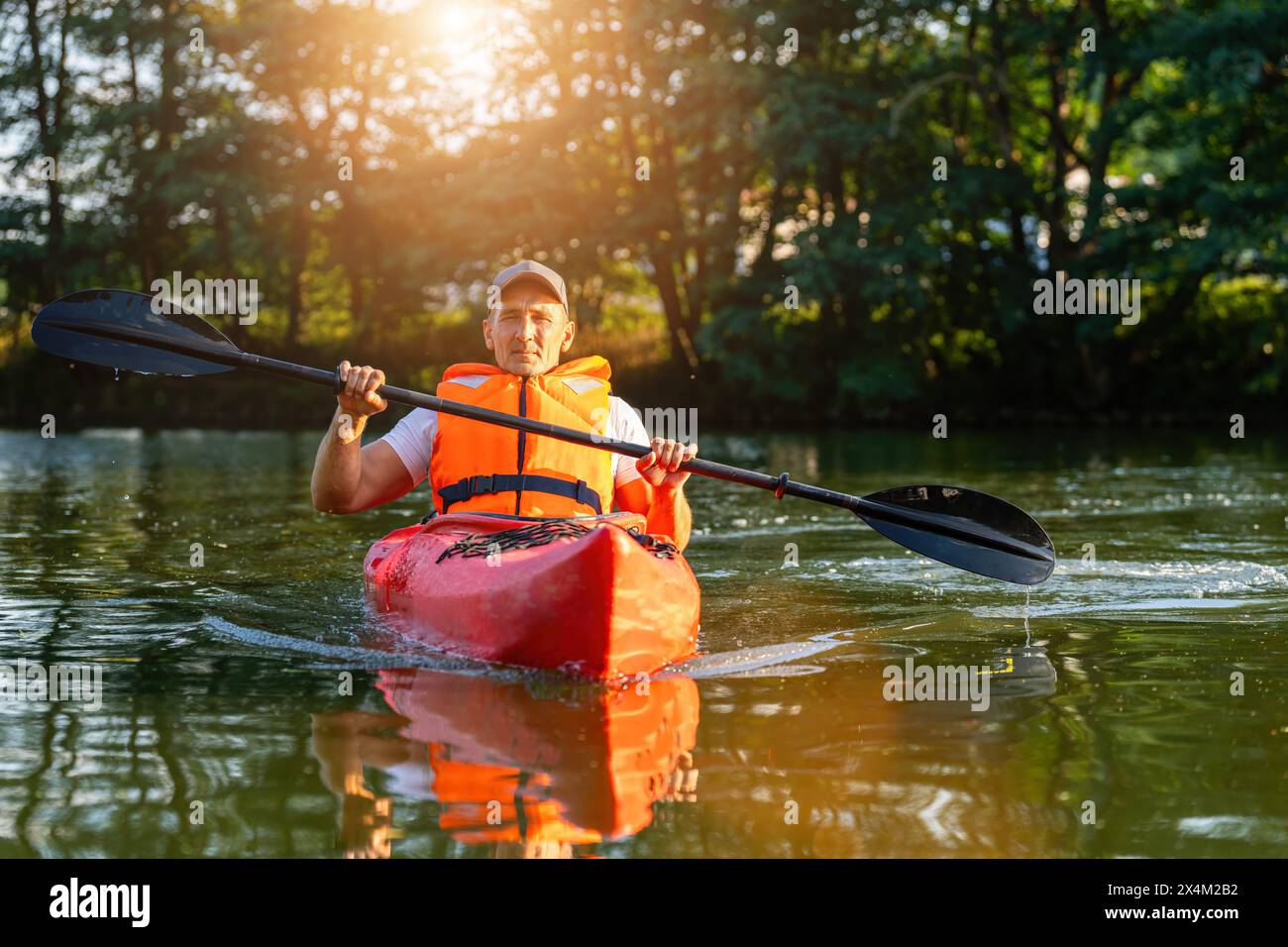 Uomo maturo con giubbotto di salvataggio arancione in kayak su un fiume tranquillo, alberi e luce del sole sullo sfondo. Immagine del concetto di kayak Water Sports Foto Stock