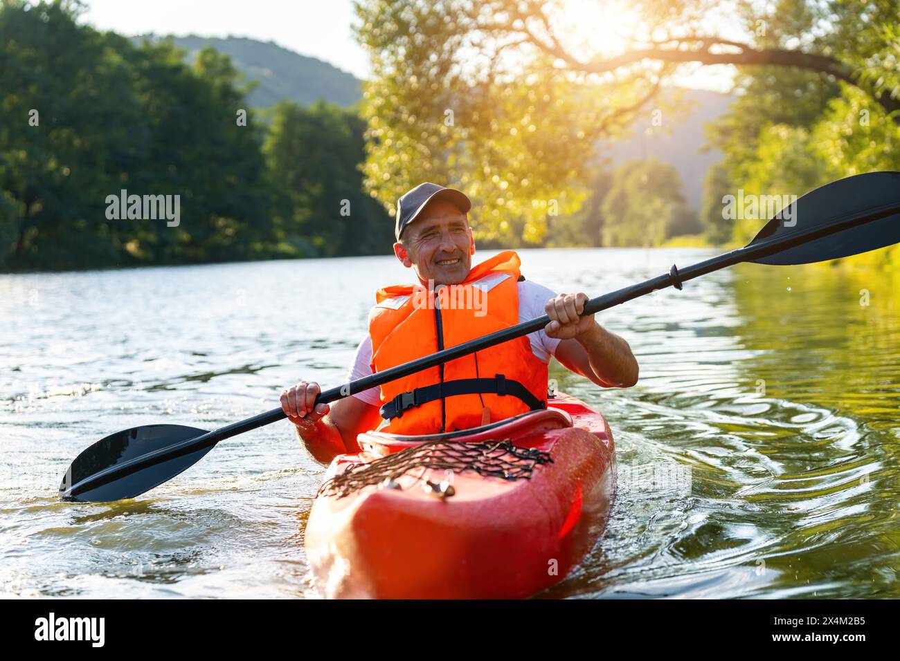 Gioioso anziano in un giubbotto di salvataggio arancione che rema un kayak rosso su un fiume, circondato da una vegetazione lussureggiante. Immagine del concetto di kayak Water Sports Foto Stock