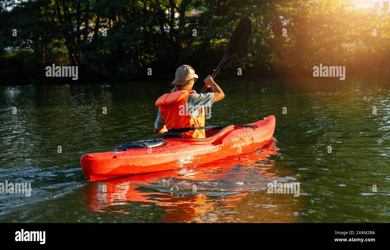 Donna con giubbotto di salvataggio arancione in kayak su un fiume tranquillo con vegetazione lussureggiante al tramonto. Immagine del concetto di kayak Water Sports Foto Stock