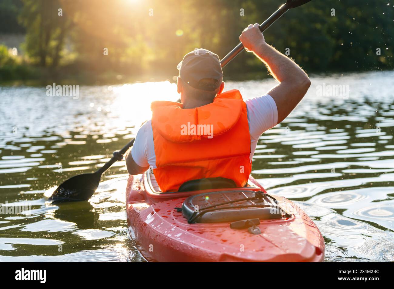 Uomo con giubbotto salvagente arancione che rema un kayak rosso su un fiume scintillante durante il tramonto in estate in germania. Immagine del concetto di kayak Water Sports Foto Stock