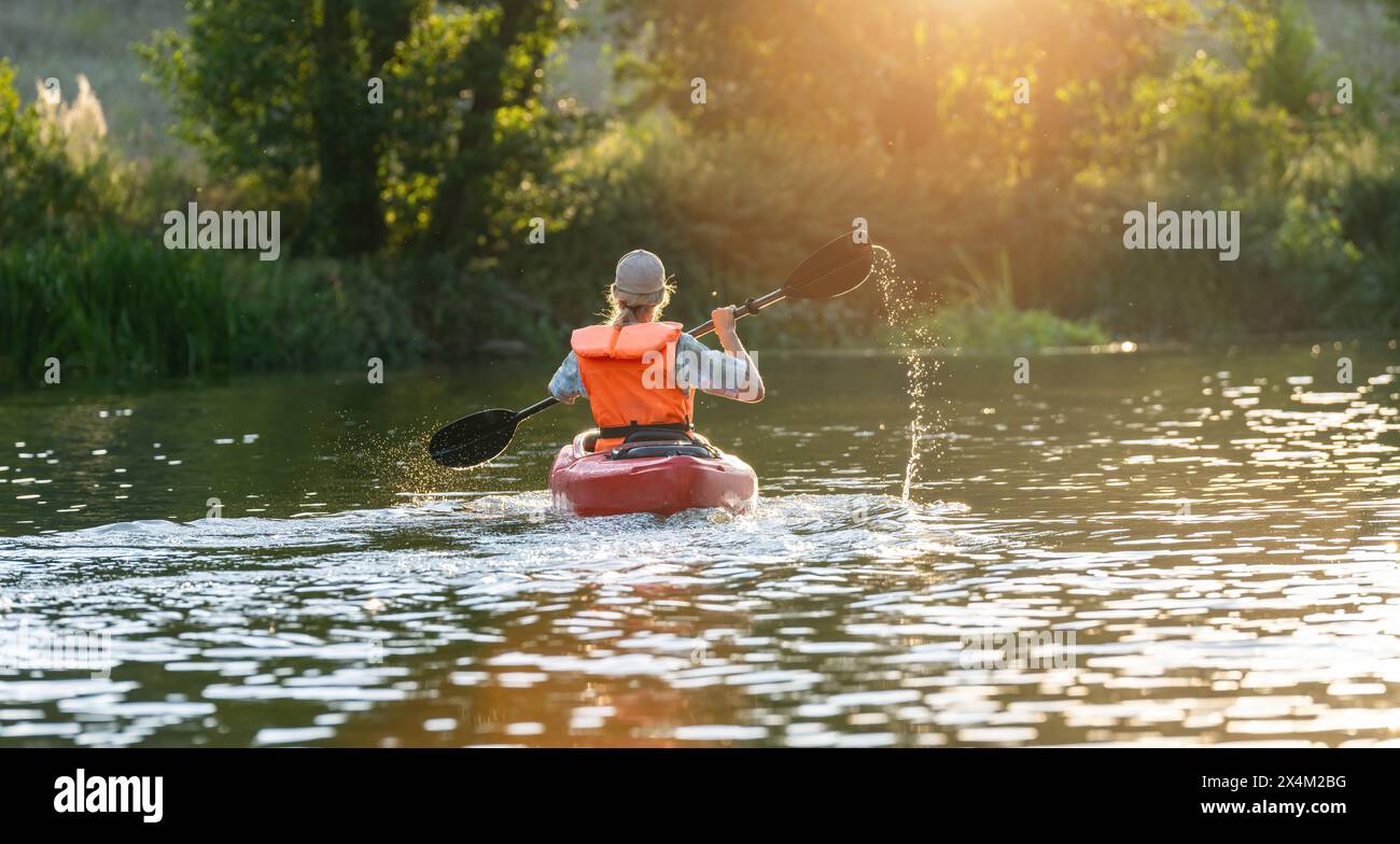 Vista posteriore di una donna che pagaia su un fiume in kayak, circondata da vegetazione lussureggiante e bagnata dalla luce del tramonto. Immagine del concetto di kayak Water Sports Foto Stock
