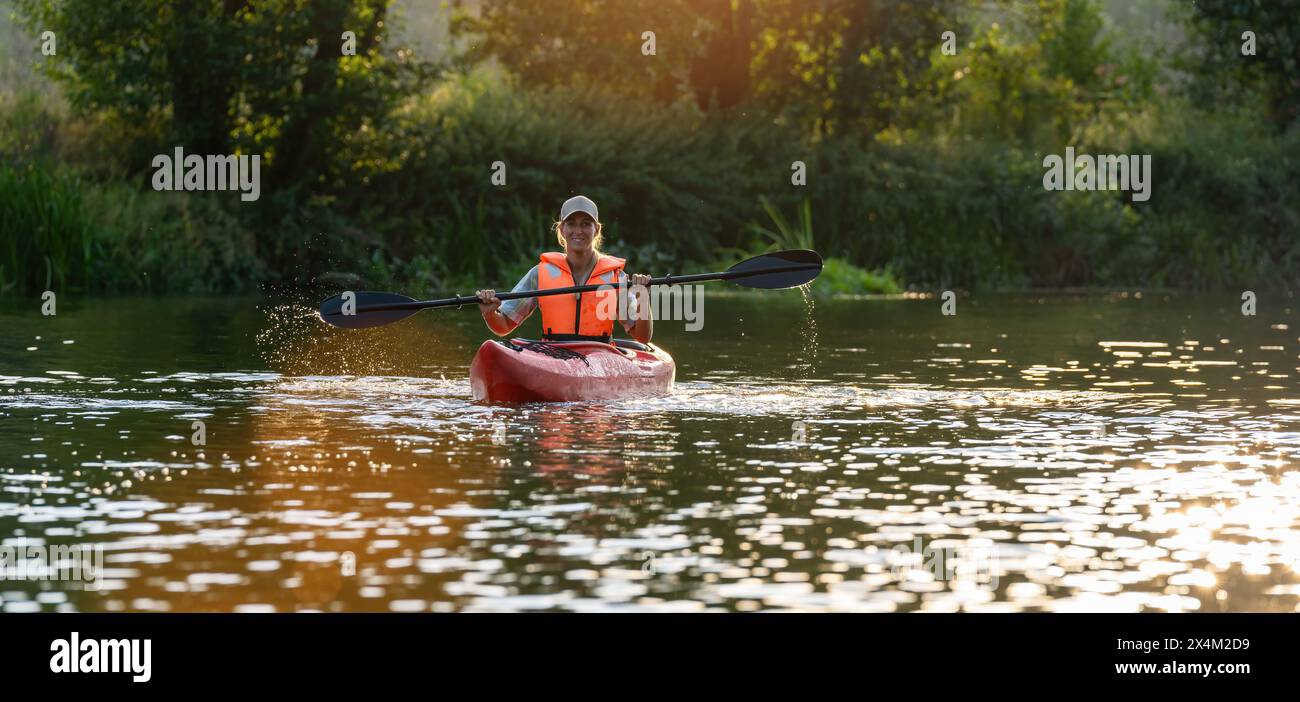 donna con giubbotto di salvataggio arancione, gioiosamente in kayak su un fiume tranquillo al tramonto, circondato da una vegetazione lussureggiante in estate in germania. Concetto di kayak per sport acquatici Foto Stock