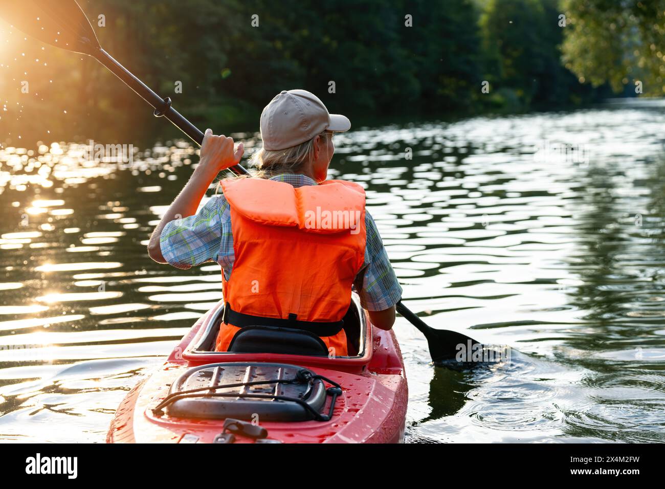 vista da dietro di una donna in un giubbotto di salvataggio arancione che rema un kayak rosso su un fiume, alberi illuminati dal sole sullo sfondo durante l'estate in germania. Kayak Water S Foto Stock