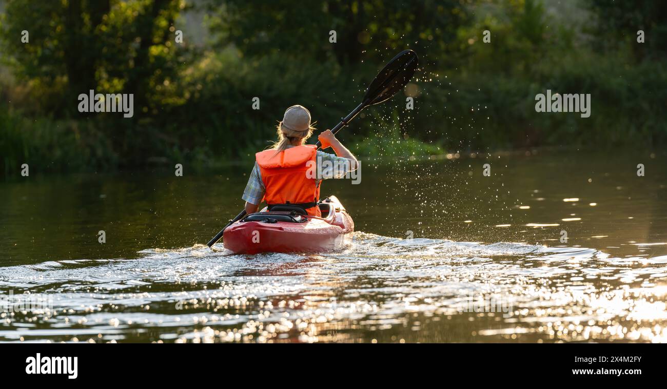 una donna in un giubbotto di salvataggio arancione rema un kayak rosso su un fiume illuminato dal sole, visto da dietro con goccioline d'acqua in aria. Immagine del concetto di kayak Water Sports Foto Stock