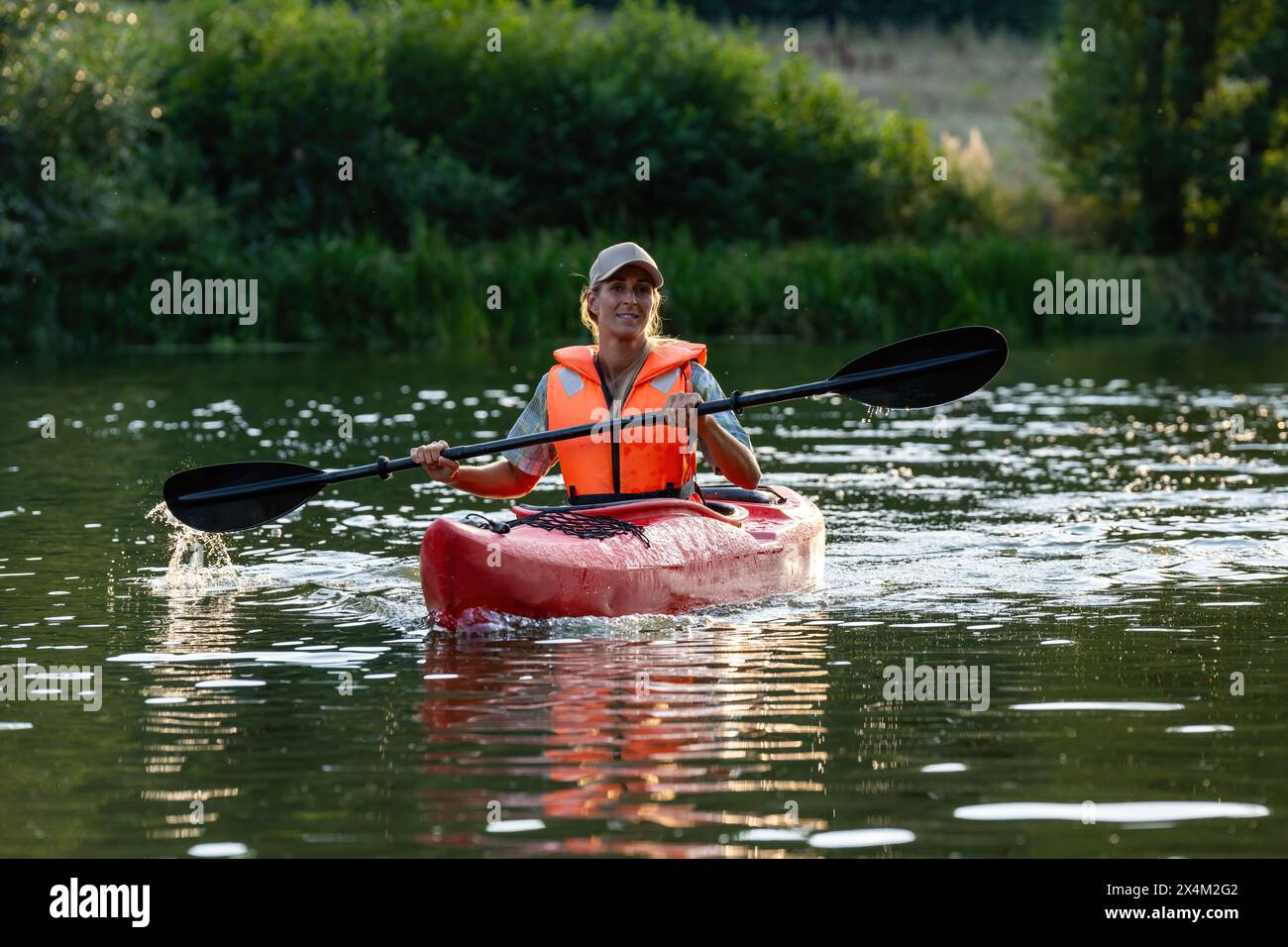 donna sorridente in kayak su un fiume calmo, remando con un reo nero, indossando un giubbotto salvagente arancione e un berretto in germania in estate. Kayak sport acquatici conc Foto Stock