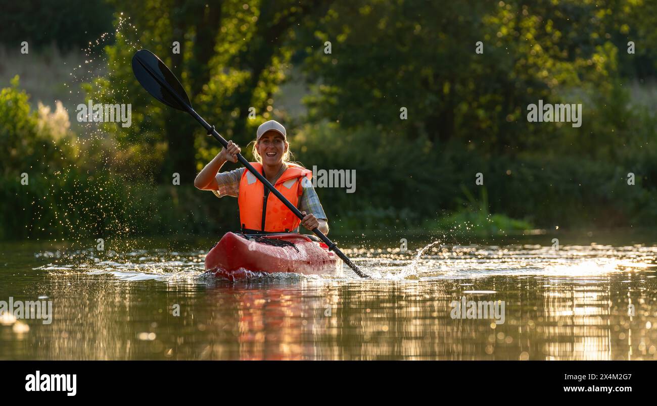 Donna felice in kayak in una giornata di sole, spruzzando acqua con la pagaia, indossando un giubbotto salvagente arancione in estate. Immagine del concetto di kayak Water Sports Foto Stock