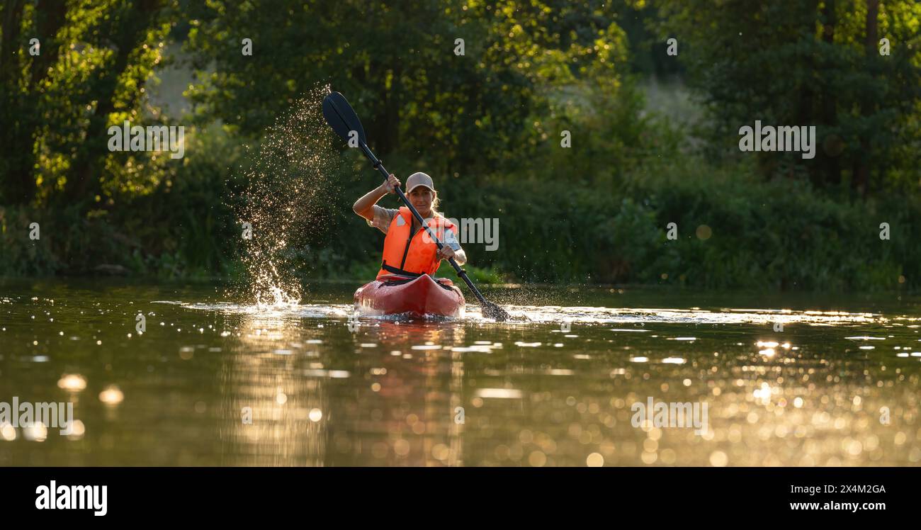 Donna in kayak nelle calme acque del fiume al tramonto, creando un tuffo con la sua pagaia che indossa un giubbotto salvagente arancione in estate. Kayak Sport acquatici concetto i Foto Stock