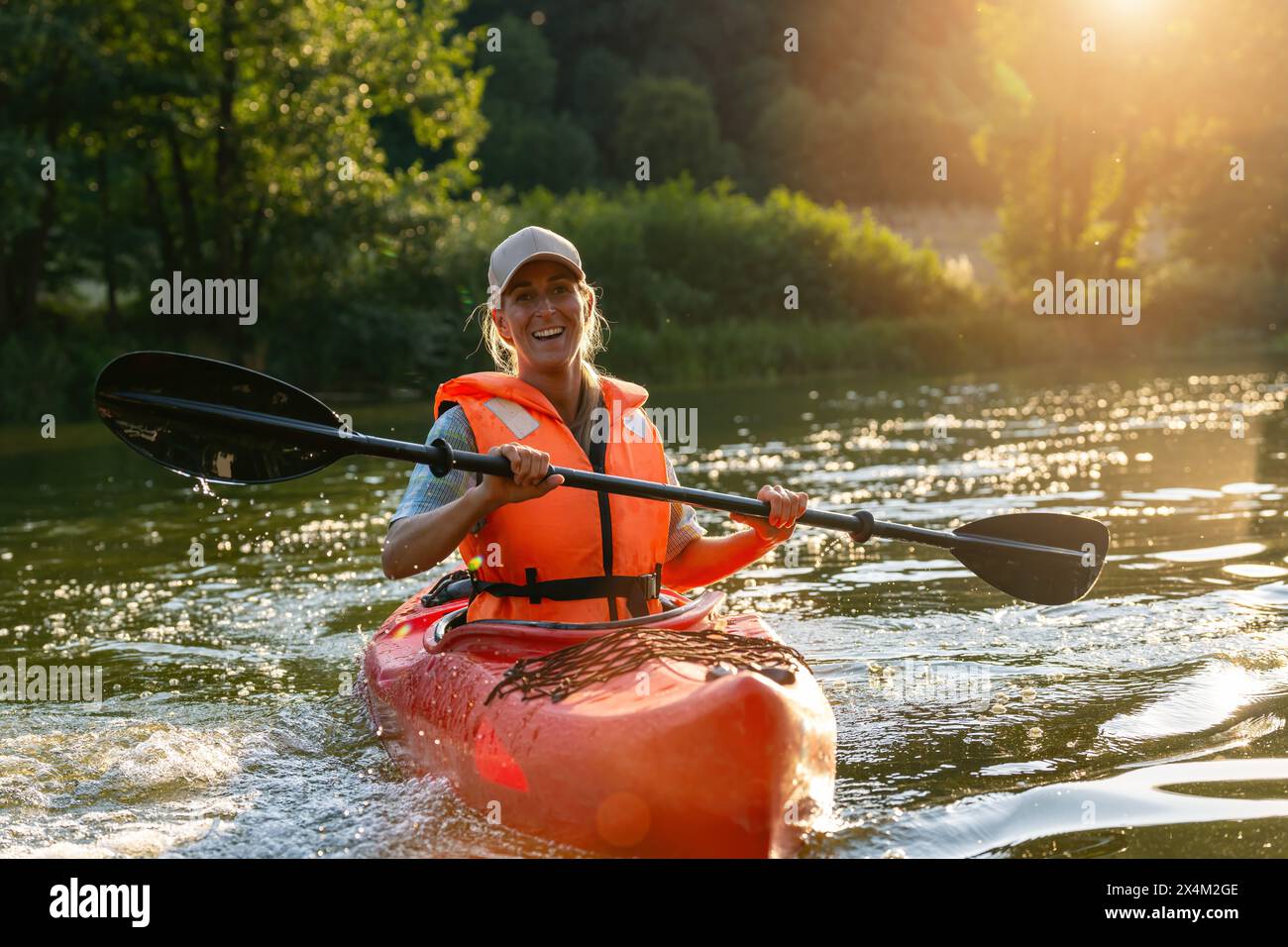 Donna gioiosamente pagaiata su un kayak rosso in un fiume durante il tramonto, circondata da una vegetazione lussureggiante. Immagine del concetto di kayak Water Sports Foto Stock
