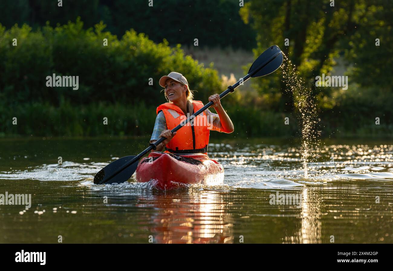 Donna sorridente che pagaiava un kayak su un fiume sereno durante l'ora d'oro. Immagine del concetto di kayak Water Sports Foto Stock