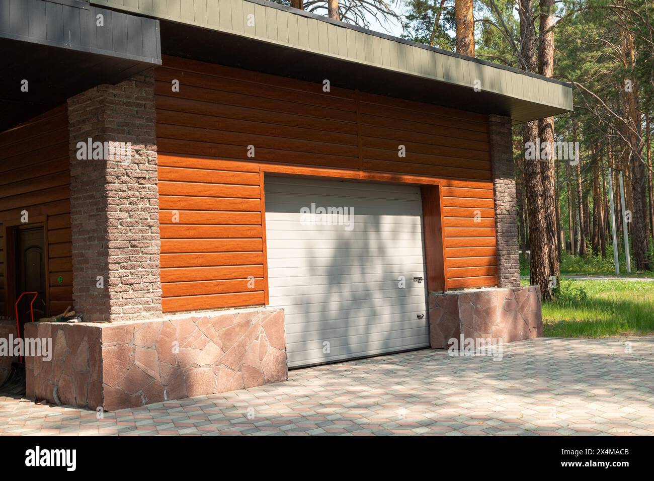 Un garage rustico con una porta bianca e un rivestimento marrone in legno in un tranquillo ambiente forestale. Le fondamenta in pietra aggiungono un fascino naturale, mentre la porta del garage chiusa migliora Foto Stock