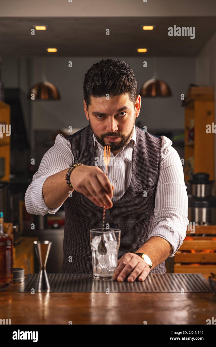 Un esperto barista maschile si concentra intensamente mentre mescola un drink presso un bancone di legno Foto Stock
