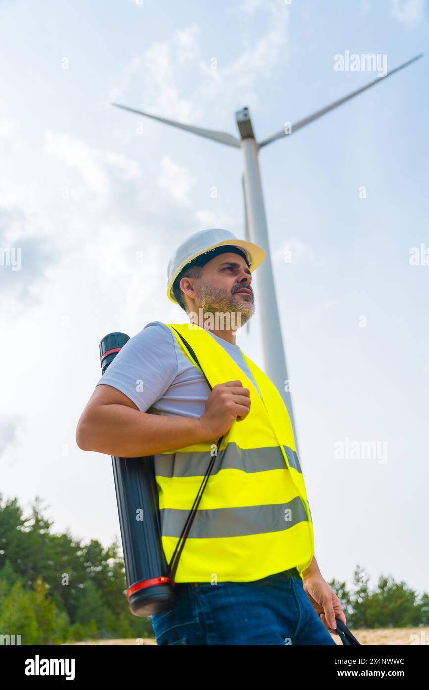 Foto verticale del profilo di un lavoratore caucasico mautre maschio con attrezzatura protettiva e tubo di disegno in un parco energetico verde accanto a una turbina eolica Foto Stock