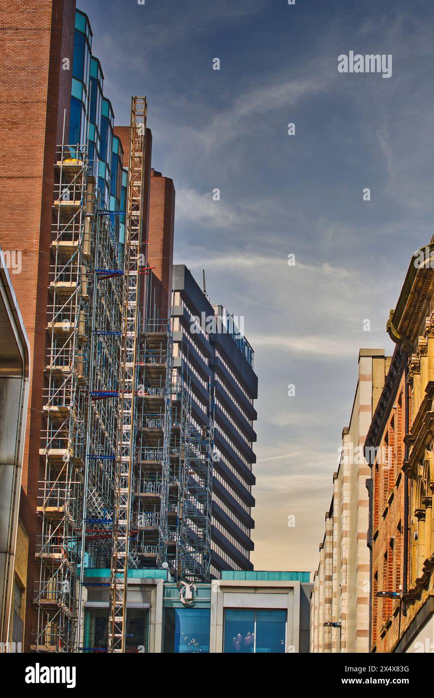 Scena urbana con una grande struttura a ponteggi su un edificio in fase di ristrutturazione, affiancato da un'architettura moderna e tradizionale sotto un cielo limpido. Foto Stock