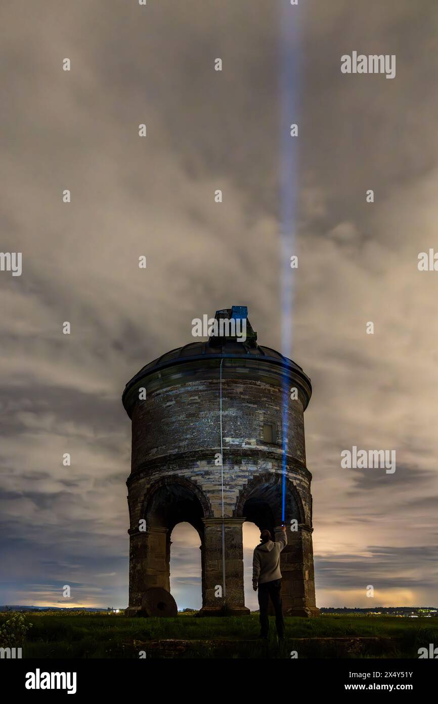 La figura solitaria si trova davanti a un antico mulino a vento, che getta un fascio di luce nel cielo notturno Foto Stock