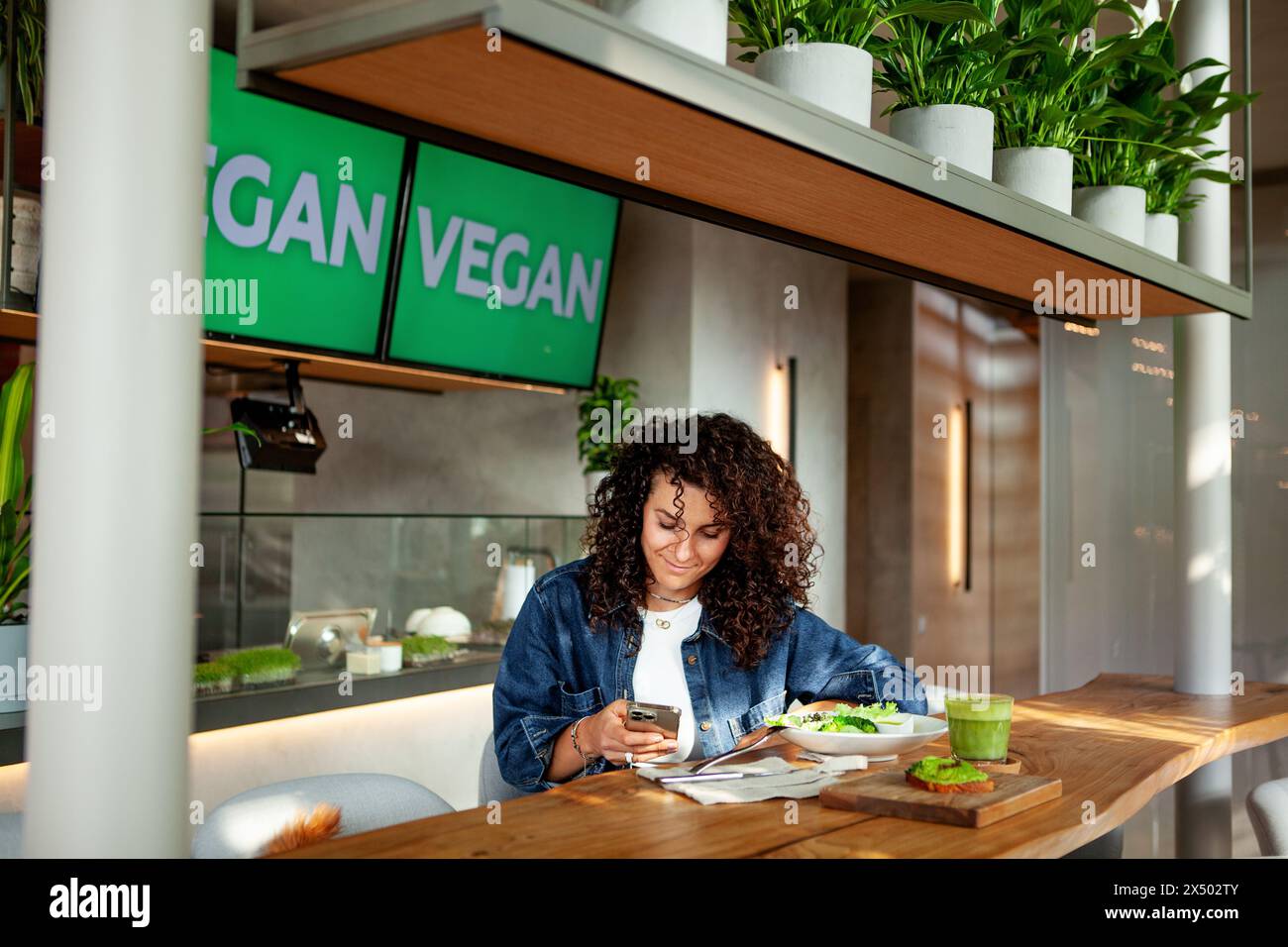 Donna con capelli ricci seduto al bar o ristorante vegetariano con frullato verde e pasto di piante sul tavolo, dall'aspetto di uno smartphone. Venues VEGAN Sign Displa Foto Stock