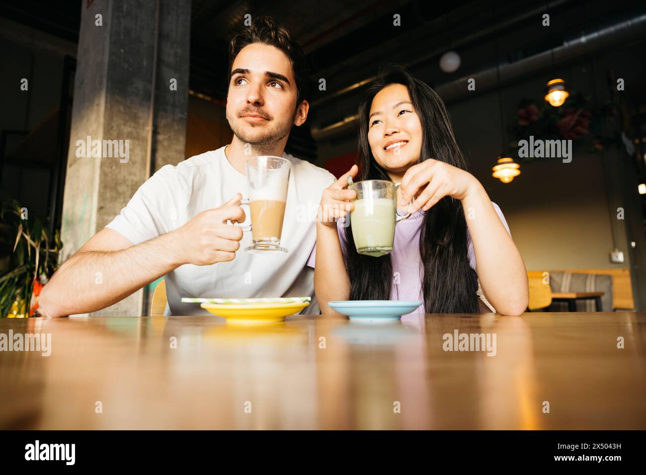 Una coppia seduta in un bar che guarda lontano e sorride. Coppia in un appuntamento in un bar locale che beve caffè e tè matcha. Foto Stock