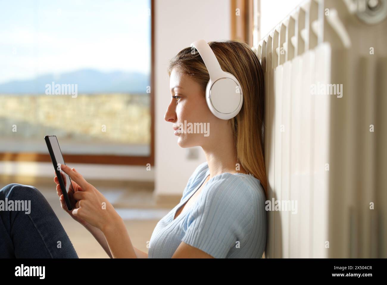 Profilo di una donna serius che si riscalda appoggiata al radiatore ascoltando musica con le cuffie e il telefono cellulare a casa Foto Stock