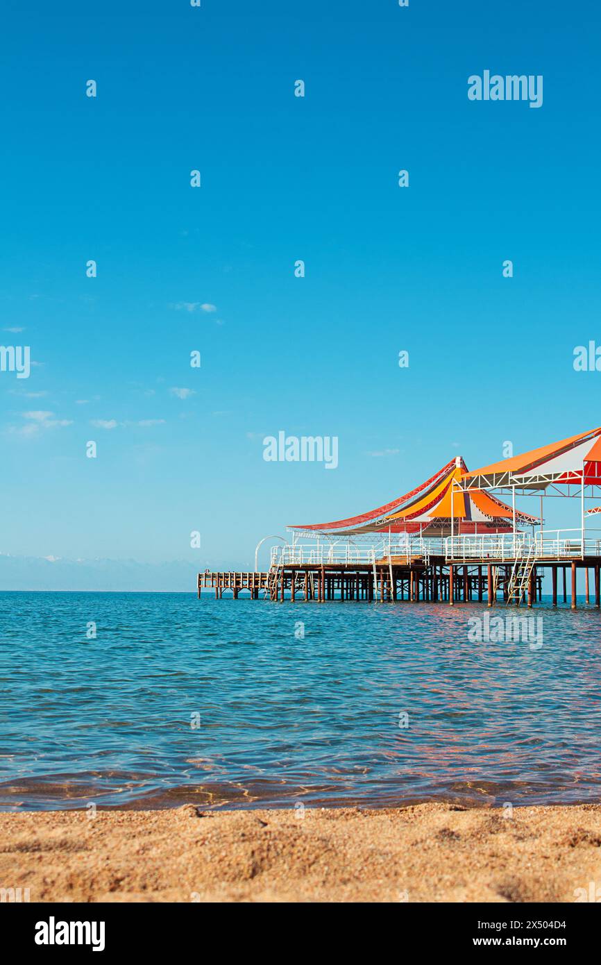 Molo in legno con tendalino rosso-arancio sull'acqua del mare, lago, oceano con spiaggia senza persone. Viaggi, vacanze, estate. Verticale. Copia spazio Foto Stock