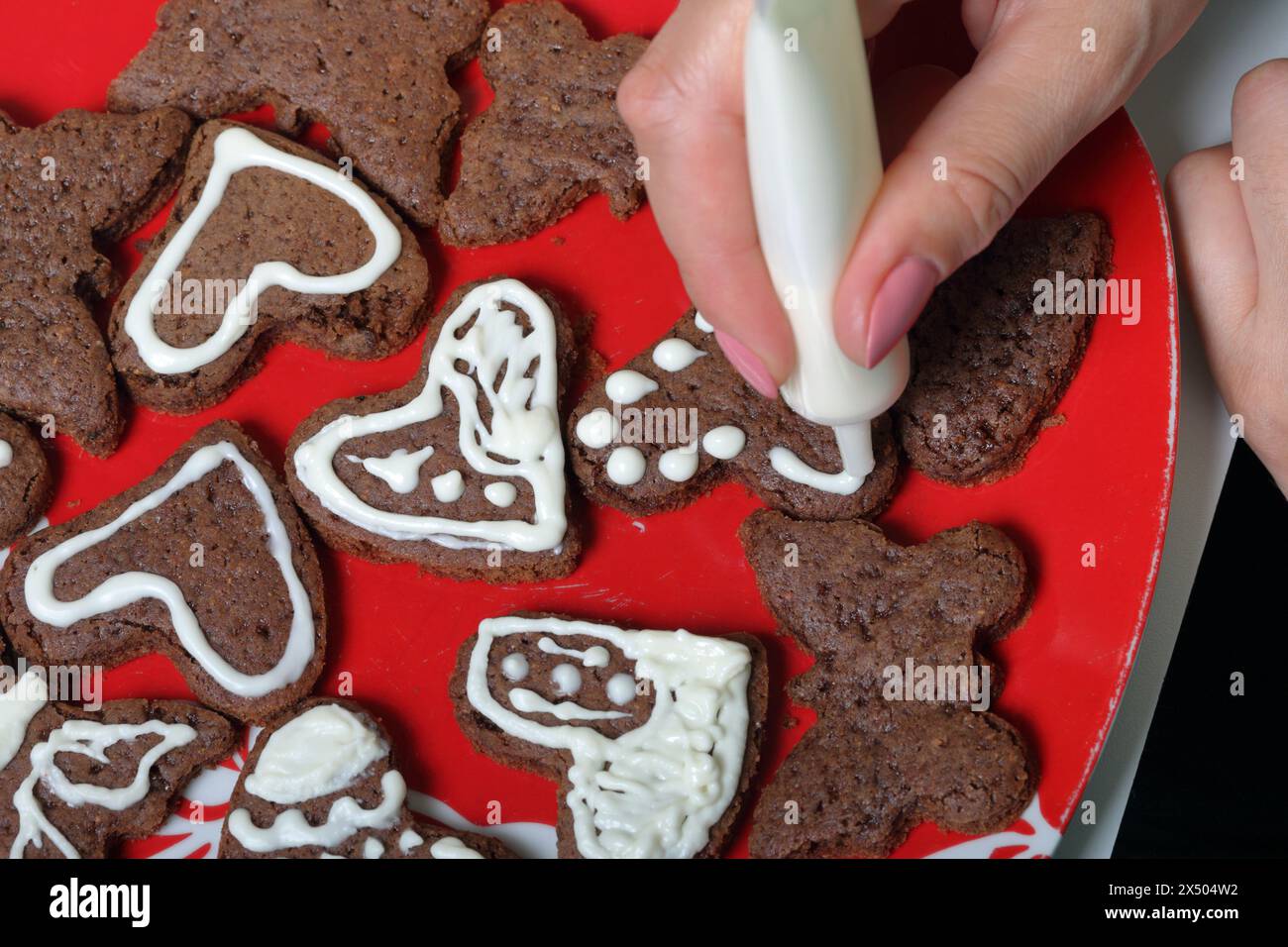 I biscotti di pan di zenzero al forno sono stesi su un piatto. Una donna spreme la glassa dal tubo, decorando i biscotti. Alcune sono decorate con ghiaccio bianco. COO Foto Stock