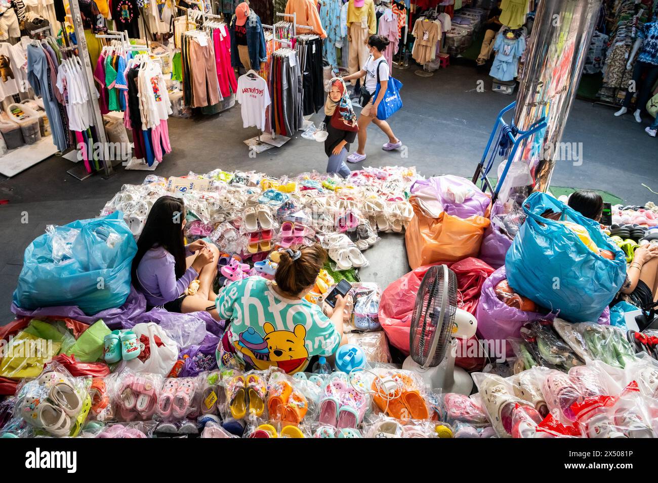 I detentori di stalle del mercato siedono tra gli abiti e i prodotti in vendita sul mercato di Pratunam. Bangkok, Thailandia. Foto Stock