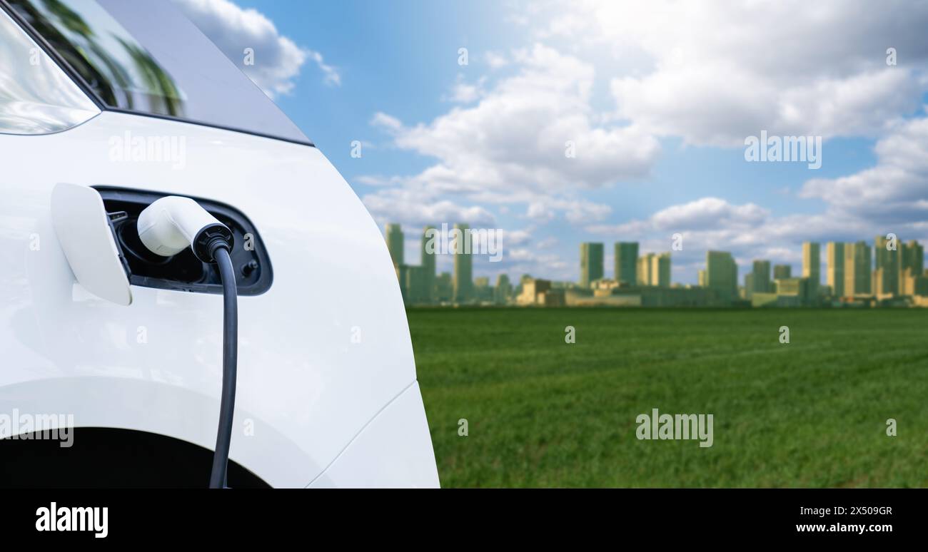 Primo piano di un'auto elettrica con un cavo di ricarica collegato sullo sfondo di una città ecologica verde. Foto Stock