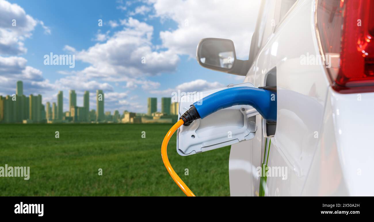 Primo piano di un'auto elettrica con un cavo di ricarica collegato sullo sfondo di una città ecologica verde. Foto Stock
