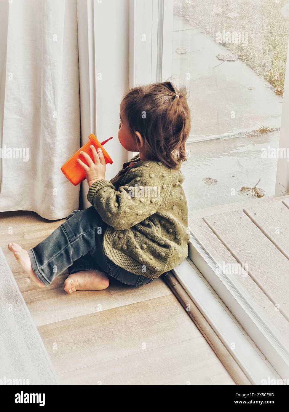 Bambino seduto su un pavimento di legno vicino a una porta di vetro che regala un drink Foto Stock