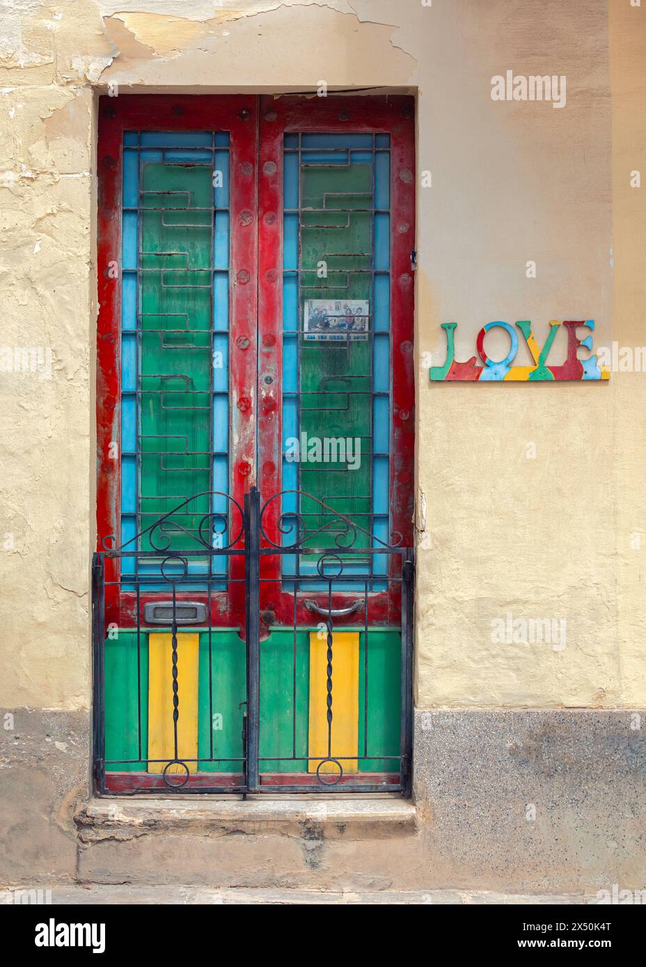 Una porta maltese vibrante e intemprata, adornata da pannelli colorati contro una rustica parete gialla. Sul muro accanto c'è un cartello che dice "amore". Foto Stock