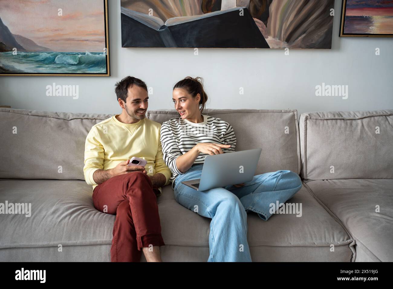 Una donna interessata mostra un uomo felice sullo schermo di un notebook, si siede sul divano, parla, fa shopping online a casa. Foto Stock