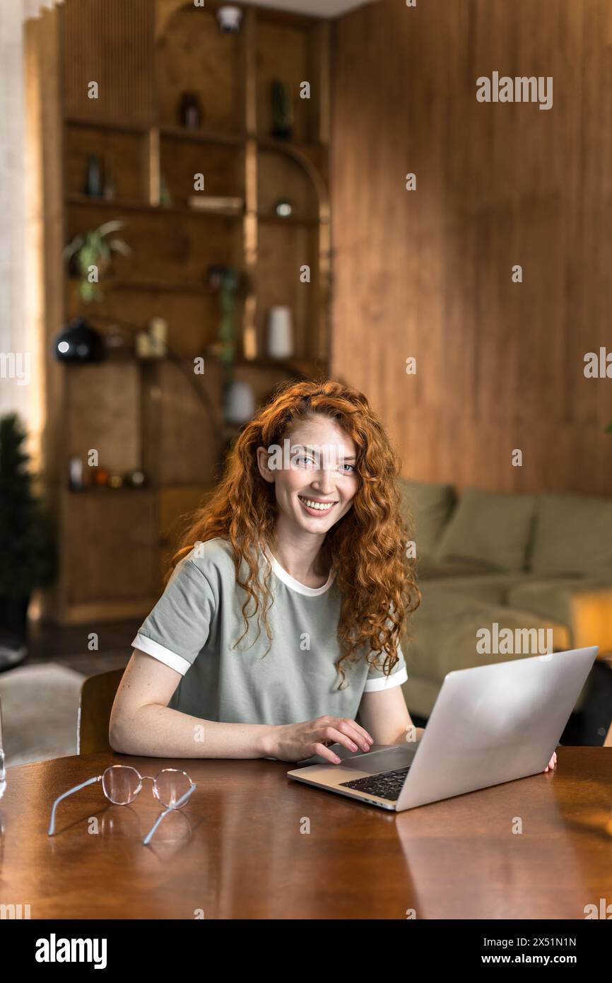 una donna dai capelli rossi sorridente siede a un tavolo e lavora a casa con un notebook Foto Stock