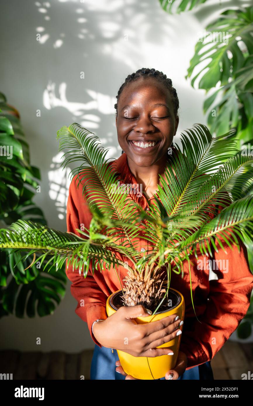 Donna nera felice che abbraccia la pentola con la pianta Cycas. Gioiosa ragazza afroamericana sorridente al negozio di fiori Foto Stock