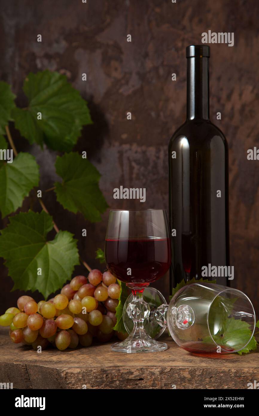 Il concetto di una deliziosa bevanda alcolica, vino e uva. Bottiglia di vino, mazzo di uva, bicchiere vuoto e bicchiere di vino rosso su legno rustico scuro Foto Stock