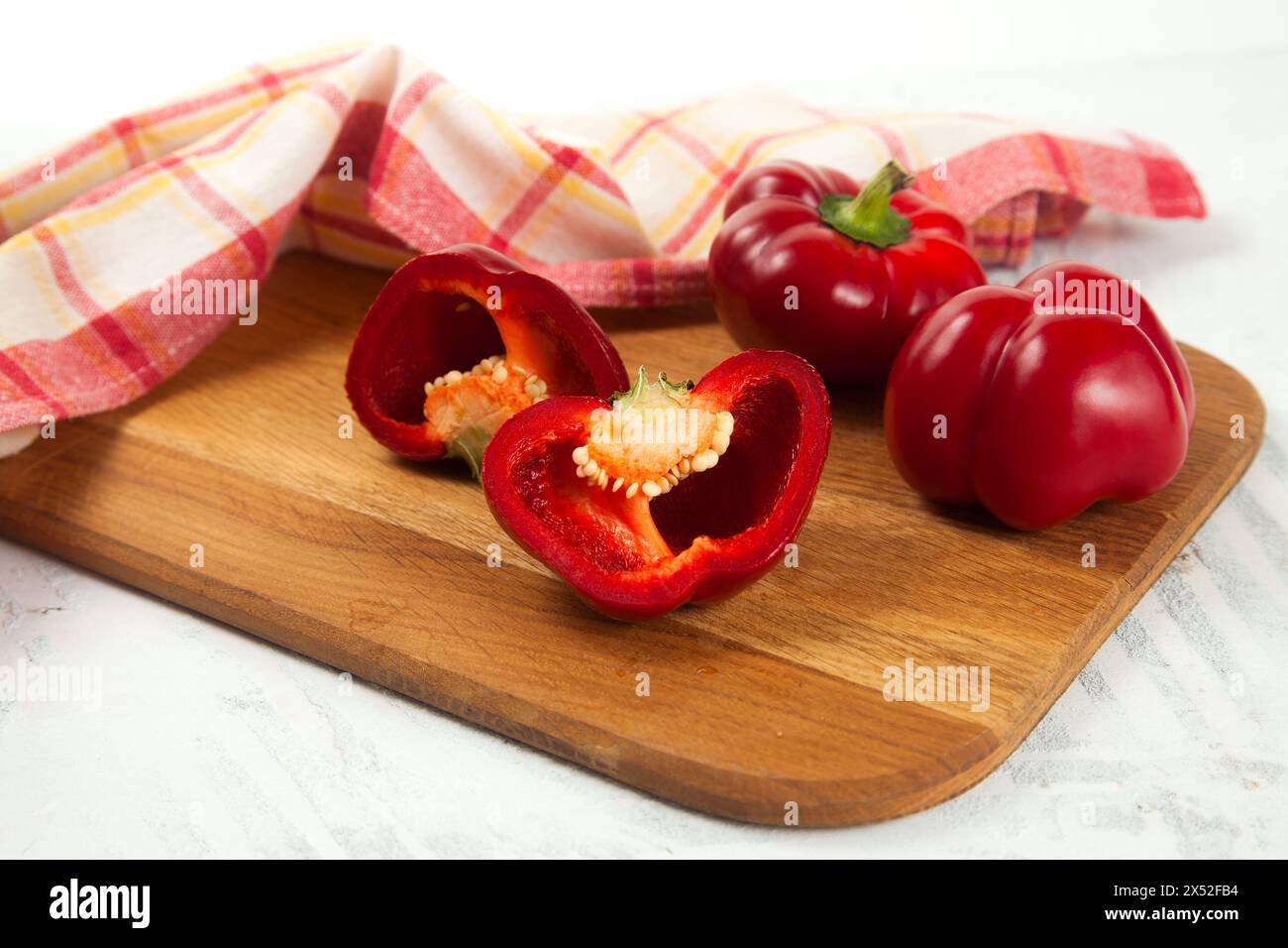 Tagliere con peperone rosso intero e mezzo (capsicum) noto come peperone dolce, paprika e asciugamano da cucina rosso su fondo di legno bianco. Foto Stock