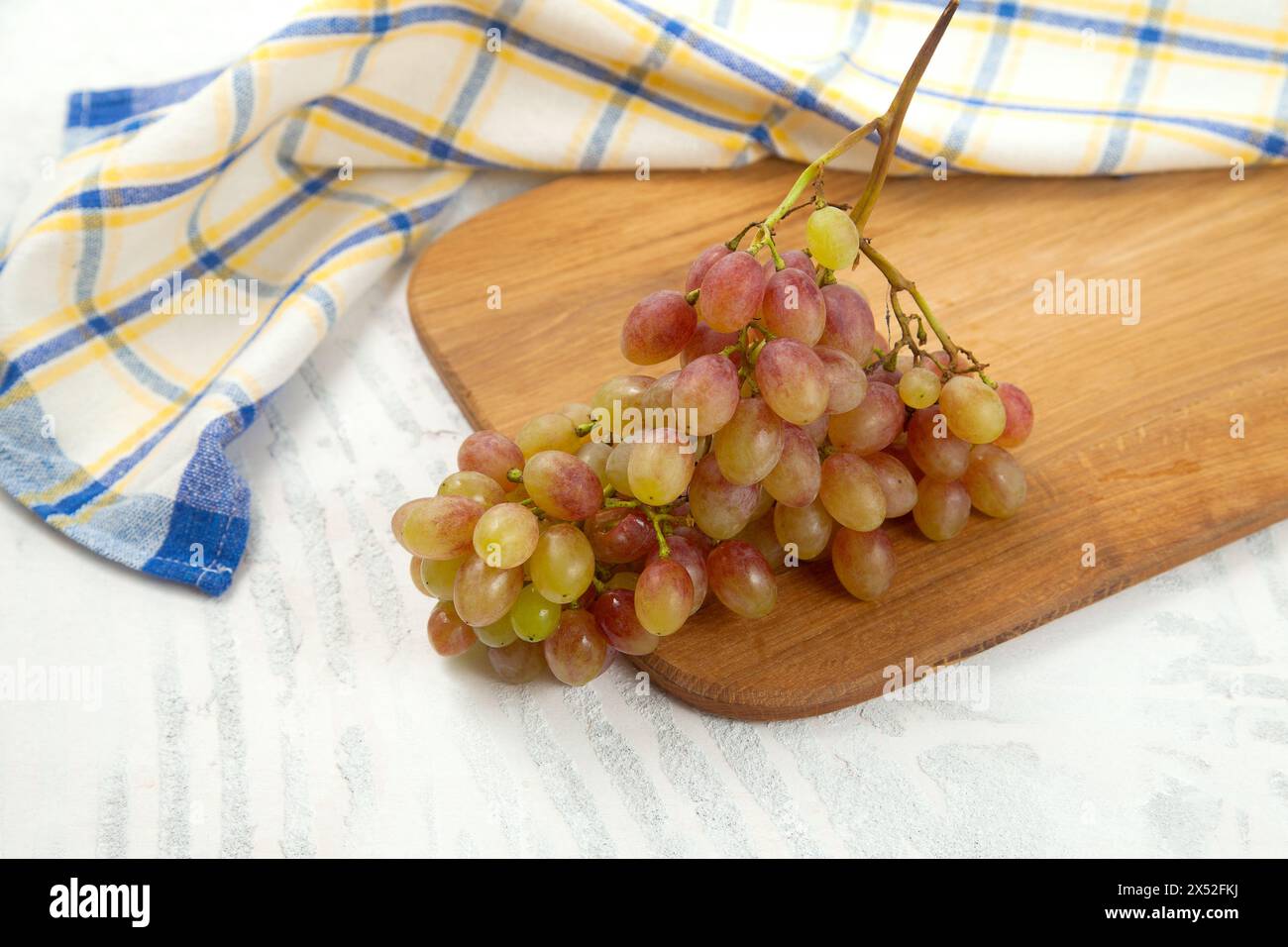 Tagliere con grande mazzo di uva rosa matura su bianco. Uva da vino rosa con asciugamano da cucina blu su fondo di legno bianco. Foto Stock