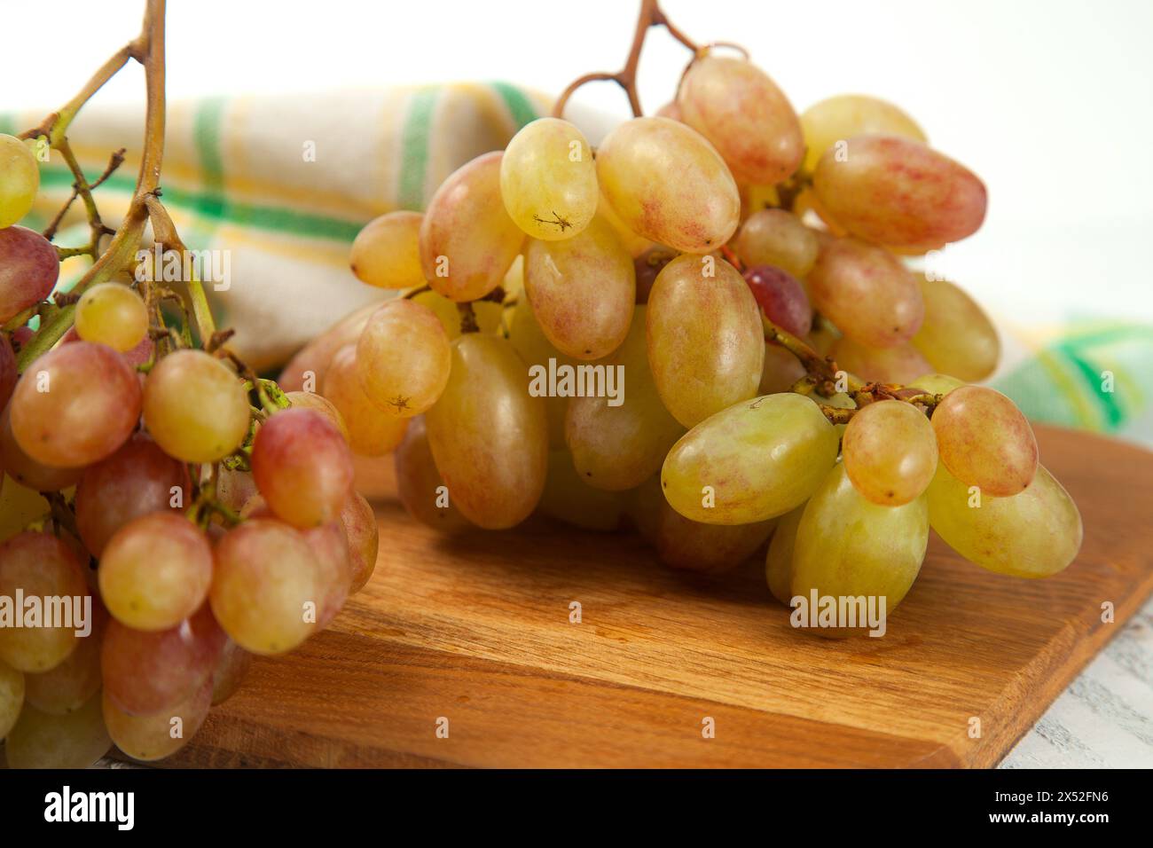 Vista ravvicinata di grandi grappoli di uva rosa matura su un tagliere di legno. Uva da vino rosa con telo da cucina verde su fondo di legno bianco. Foto Stock