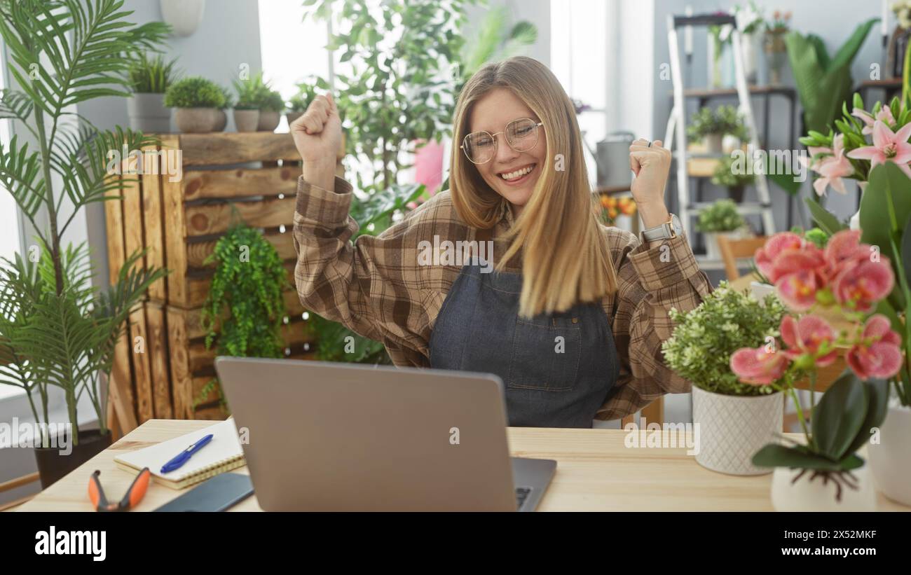 Una giovane donna gioiosa con capelli biondi celebra il successo in un vivace negozio di fiori, circondato da vegetazione e fiori. Foto Stock