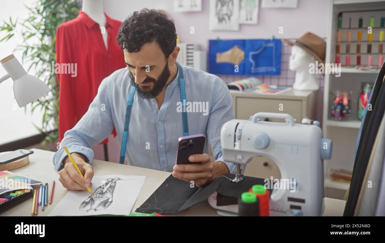 Un uomo affascinante sartoriale in un atelier moderno, che usa il telefono mentre disegna un design di moda. Foto Stock