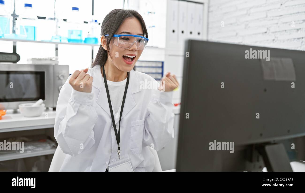 Una donna asiatica entusiasta scienziata in un laboratorio che indossa occhiali di sicurezza e un camice da laboratorio celebra una svolta guardando uno schermo di un computer. Foto Stock