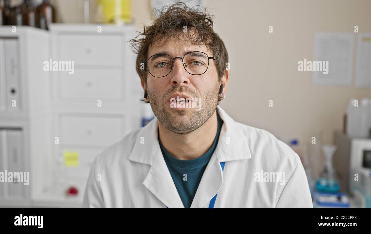 Ritratto di un giovane uomo ispanico barbuto che indossa un camice da laboratorio e occhiali da vista al chiuso in una clinica medica. Foto Stock