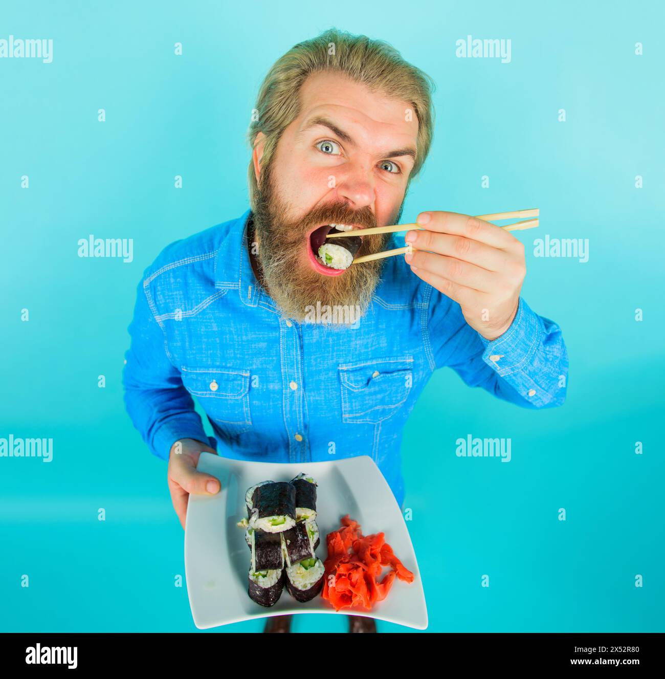 Cucina tradizionale giapponese. Bell'uomo in camicia di denim che mangia sushi roll con bacchette. Cibo di mare. Elegante uomo barbuto con piatto di deliziosi maki Foto Stock